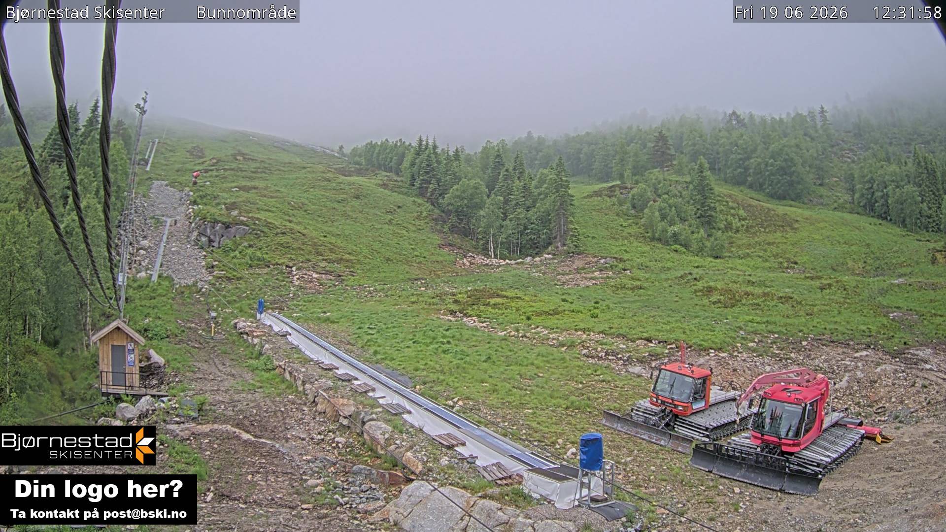 A foggy summer ski slope: bare green hillside, low cloud over the ridge, two red piste groomers parked and idle at the base, a small wooden lift hut, a conveyor lift, scattered stone.