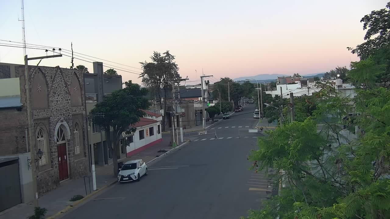 A small-town Argentine street at dawn — pink-orange sky, a stone church with gothic arches, one parked white car, empty road, the first golden light.