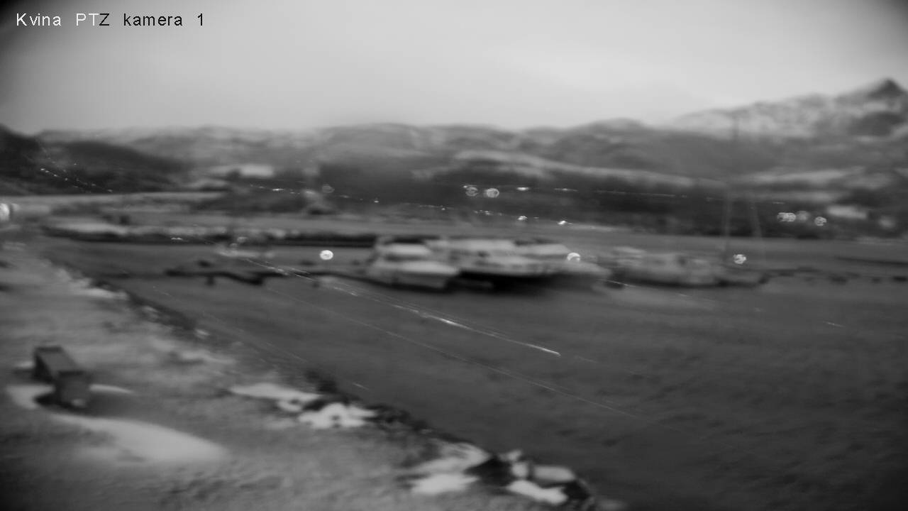 Black-and-white dusk webcam view of Kvina harbour, Norway — snow-dusted mountains, moored boats, flat grey winter light, snow patches in the foreground.
