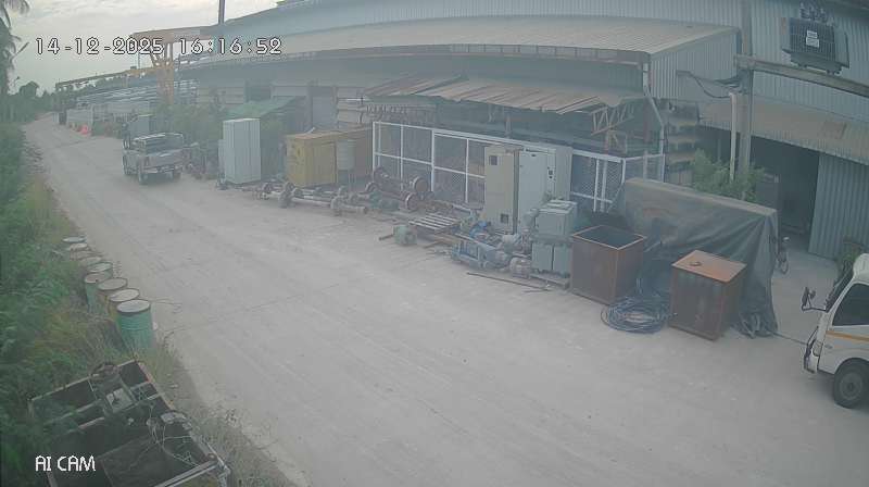 An industrial yard in Thailand in flat afternoon light — a warehouse with a curved metal roof, barrels and pipes along the wall, a parked pickup truck, the label AI CAM in the corner.