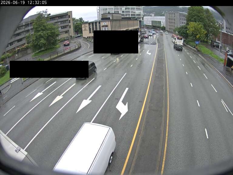 A wet multi-lane city road seen through a fisheye lens, double yellow centre line, painted lane arrows, a white van in the foreground, pale buildings and green hills behind, with two black rectangles redacting vehicles' licence plates.